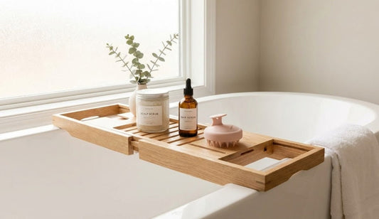 A relaxing bath setup featuring a jar of scalp scrub, a hair serum bottle, and a pastel silicone scalp massager on a wooden tray, decorated with eucalyptus leaves and a white towel.